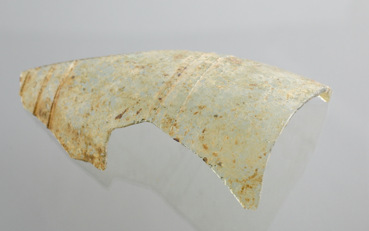 Del av buken på ett ofärgat, kägelformat, optikblåst passglas som är ornerat med fem pålagda parallella trådar. De två övre trådarna är strierade. Fragmetet har glaspest.