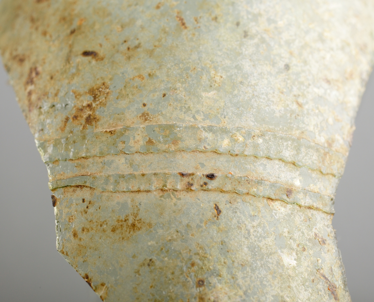 Del av buken på ett ofärgat, kägelformat, optikblåst passglas som är ornerat med fem pålagda parallella trådar. De två övre trådarna är strierade. Fragmetet har glaspest.