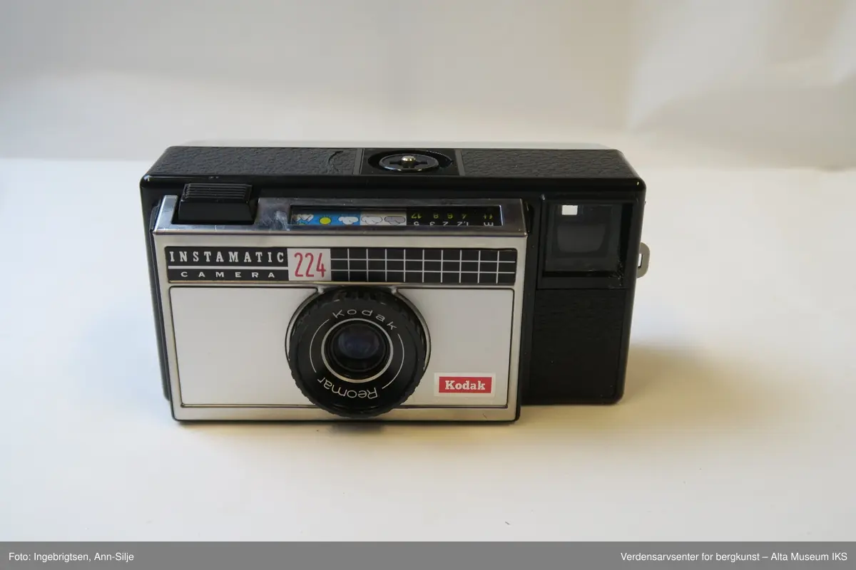 Fotoapparat i rektangulær form. Den er festet i et læretui med rødt tøystoff på innsiden. Inn i apparatet er det en filmrull av merket Fuji. På toppen av apparatet, ved siden av utløseren, er det symboler av vær og landskap.