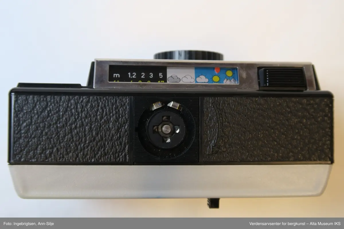 Fotoapparat i rektangulær form. Den er festet i et læretui med rødt tøystoff på innsiden. Inn i apparatet er det en filmrull av merket Fuji. På toppen av apparatet, ved siden av utløseren, er det symboler av vær og landskap.