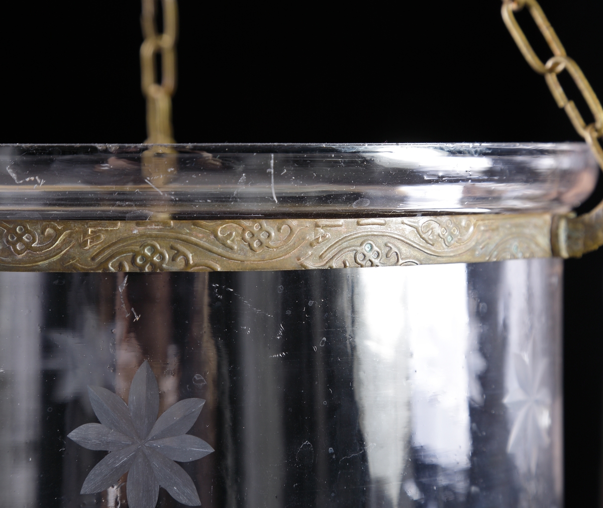 Ljusampel av glas.
Cylindrisk form med rund botten, avslutad med knopp. Dekor av slipade stjärnor (blommor?). Runt ampelns övre kant löper ett band av graverad mässing, med tre gjutna kedjehållare utformade som sjöhästar. Över ampeln ett profilerat, runt ljustak av glas. Kedjor av mässing.