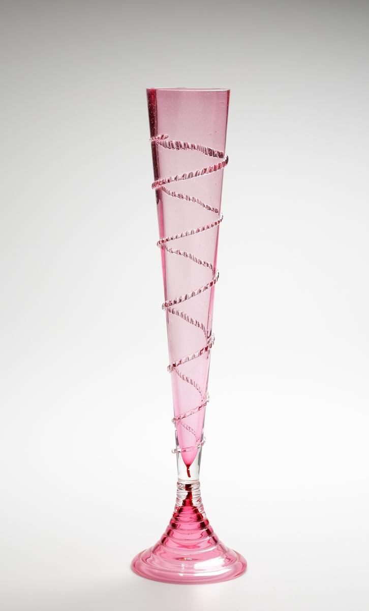 Vas. eller möjligen pokal.
Stilen på glasen är gjord så att den ska se ålderdomlig ut.
Hög rosa, konisk vas med spunnen och naggad ofärgad glastråd. 
Foten rosa  och konisk med markerade ränder.