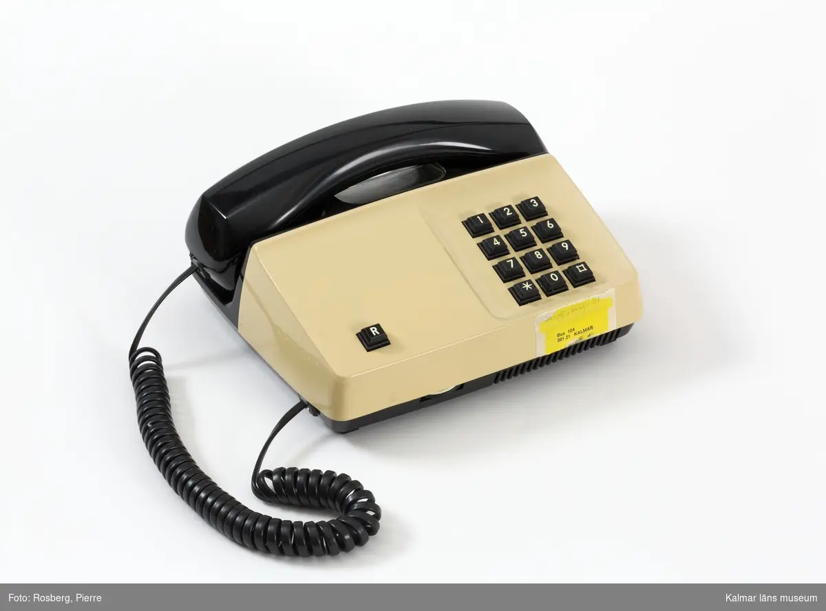 KLM 46164:1-2. Telefon, telefonapparat, knapptelefon. Av beige och svart plast. Tillverkare: Telia, Televerket. Modell: Diavox. I rektangulär plan form med svart knappsats till höger. Vid fronten en gul etikett med angiven adress. Medföljer svart elsladd och förlängningssladd med vit telefonkontakt.