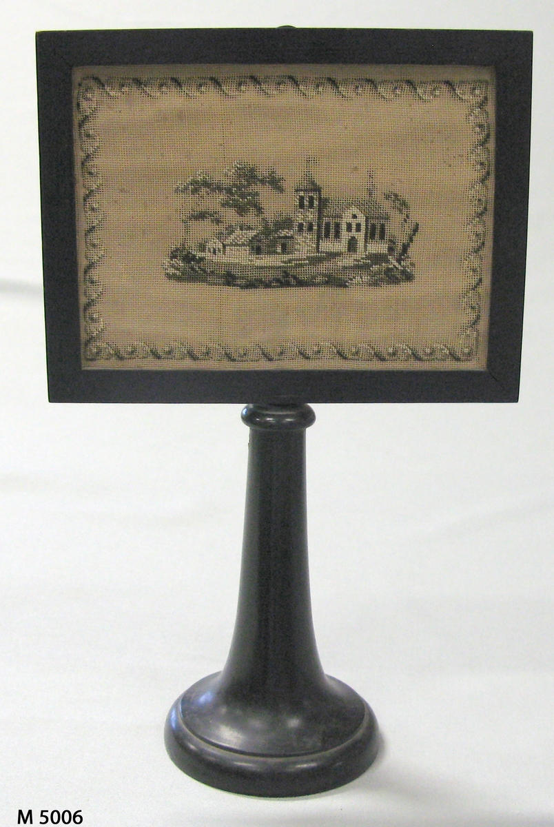 Ljusskydd.
Svart träram på svarvad träfot. I ramen körssömsbroderi å stramalj i 
färgerna gråsvart och vitt. Motiv: kyrka. Baksidan täckt med grönt siden (japonne).
Ramens storlek: 19,5 x 15 cm. Fotens höjd: 16,5 cm. 
Diameter nedtill 10 cm.
Funktion: Ljusskydd