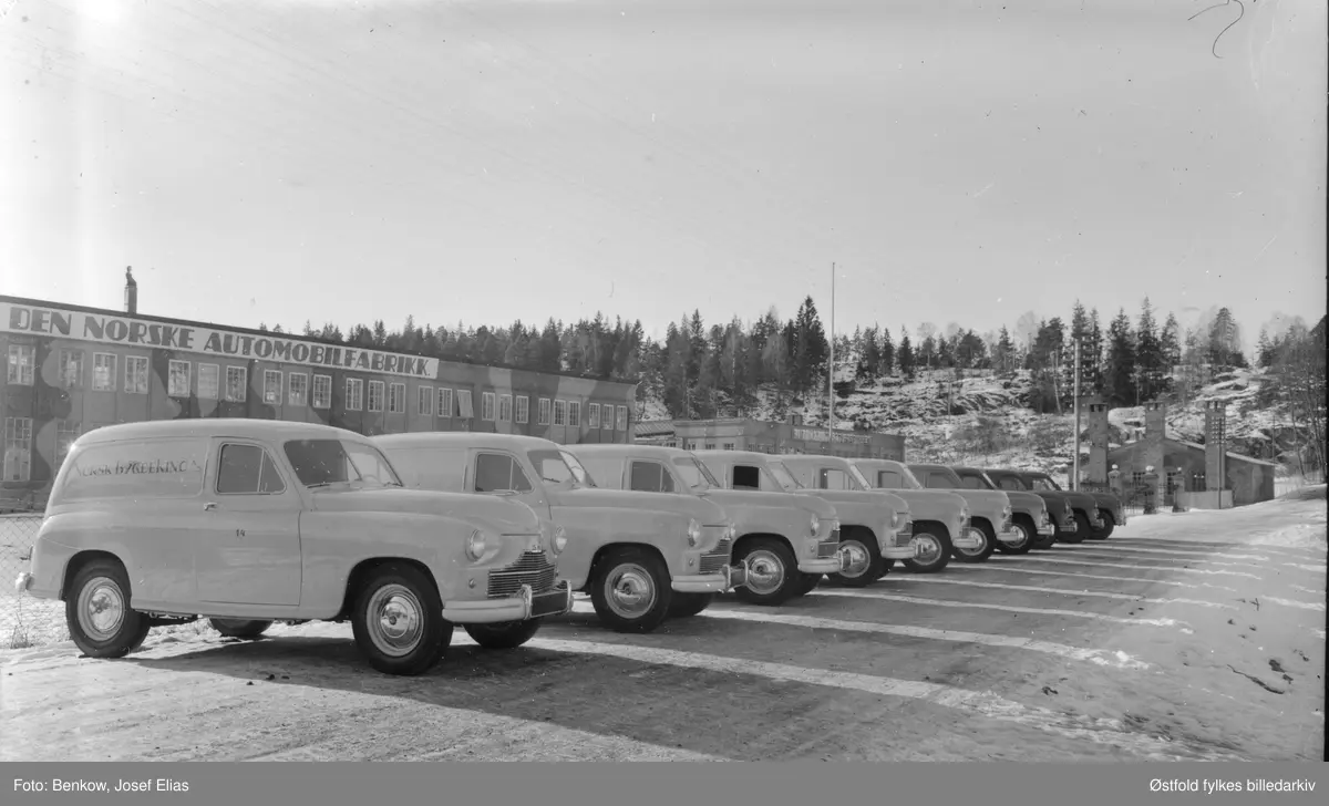 Nyproduserte biler til Norsk Bygdekino A/S utenfor Den Norske Automobilfabrikk på Kambo i Moss. Standard Vanguard  (1948-52) ble klargjort, men ikke montert på Kambo. Oppstilt rekke av  gule varevogner til Norsk Bygdekino A/S som ble etablert 23. juli 1950. 
Fabrikkbygningen har fortsatt kamuflasjemaling fra 2. verdenskrig. 
Fabrikken ble rekvirert av tyskerne under 2. verdenskrig og ble brukt som verksted for tyske Junker-fly, overhaling av flymotorer.
Moss byleksikon: 
"Norsk Automobilfabrikk, Møllebakken 4, Kambo i Moss. Etablert i 1918. Her ble det produsert 77 lastebiler i løpet av tre år. Men så gikk bedriften konkurs i 1923. Den ble reetablert 1929 av Gustav Henning. Bedriften ble startet i samarbeid med svenske Scania-Vabis. De siste to årene ble det produsert karosserier og enkelte deler. Men 13 år senere - i 1936 - ble Den Norske Automobilfabrikk etablert, i de samme lokalene. Der ble det ikke produset biler, men karosserier til Plymouthbiler og Fargo lastebiler ble satt sammen. Bedriften kunne fremstille gjennomsnitlig ett karosseri om dagen. Det var stort sett lasteplan og førerhus som ble laget på fabrikken. Den totale produksjonen ble anslått til i underkant av 2200 biler.
Etter krigen ble fabrikken brukt til å klargjøre importerte biler. Virksomheten opphørte i 1963-64.