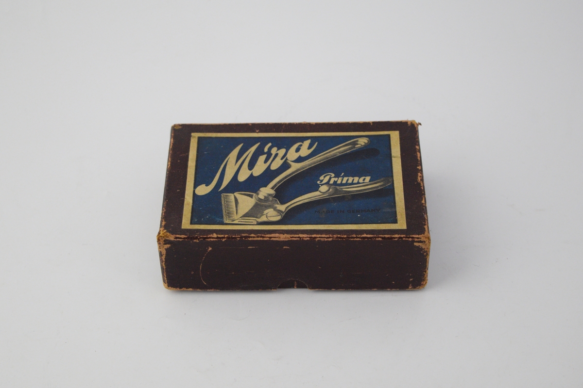 Eske inneholdende saks, tre ekstra utskiftbare deler til saksen, en fjær, en metallplate, fire pakker med flere barberblad og en lapp med instruksjoner for bruk og vedlikehold på flere språk, deriblant engelsk.