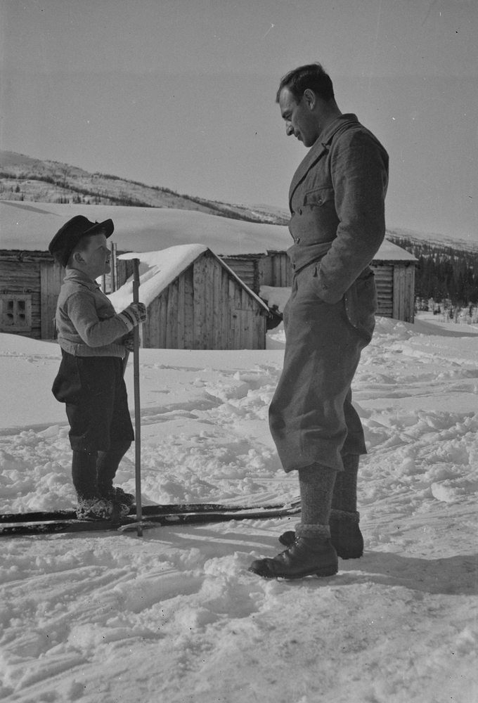 Gutt på ski som prater med en voksen mann. Gutten er Asbjørn norvald Svartvassmo.
Tømret bygning i bakgrunnen.