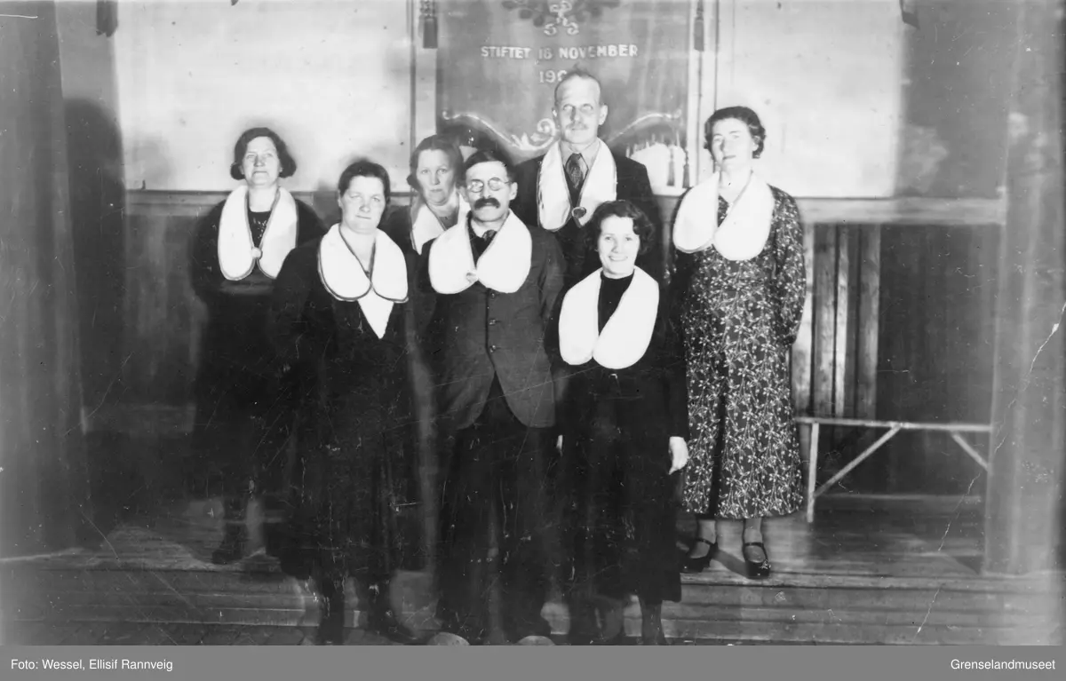 Gruppebilde av kvinner og menn, sannsynligvis foreningsmedlemmer. En fane ses i bakgrunnen, sier at foreningen/klubben er stiftet i november 190..., ukjent sted og ukjent forening.