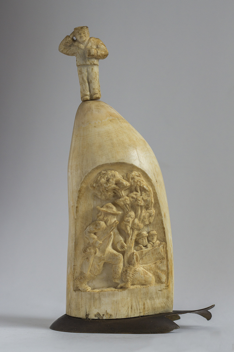 Hvaltannskulptur på plint av messing formet som en hval. Utskåret jegermotiv i hvaltannen.