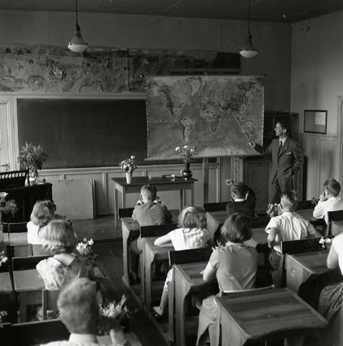 Skolavslutning, Glösbo 9 juni 1956. Lärare och elever i klassrummet. Magistern pekar på en karta och de finklädda eleverna sitter i sina skolbänkar och lyssnar. På katedern och orgel står blombuketter.