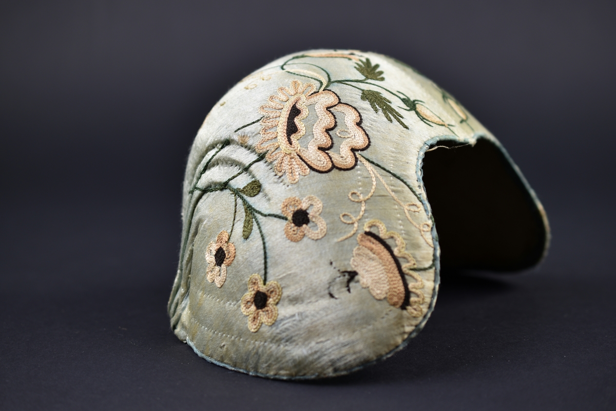 Bindmössa av ljusblått siden dekorerad med broderade blommor och blad. Foder i oblekt linnetyg.