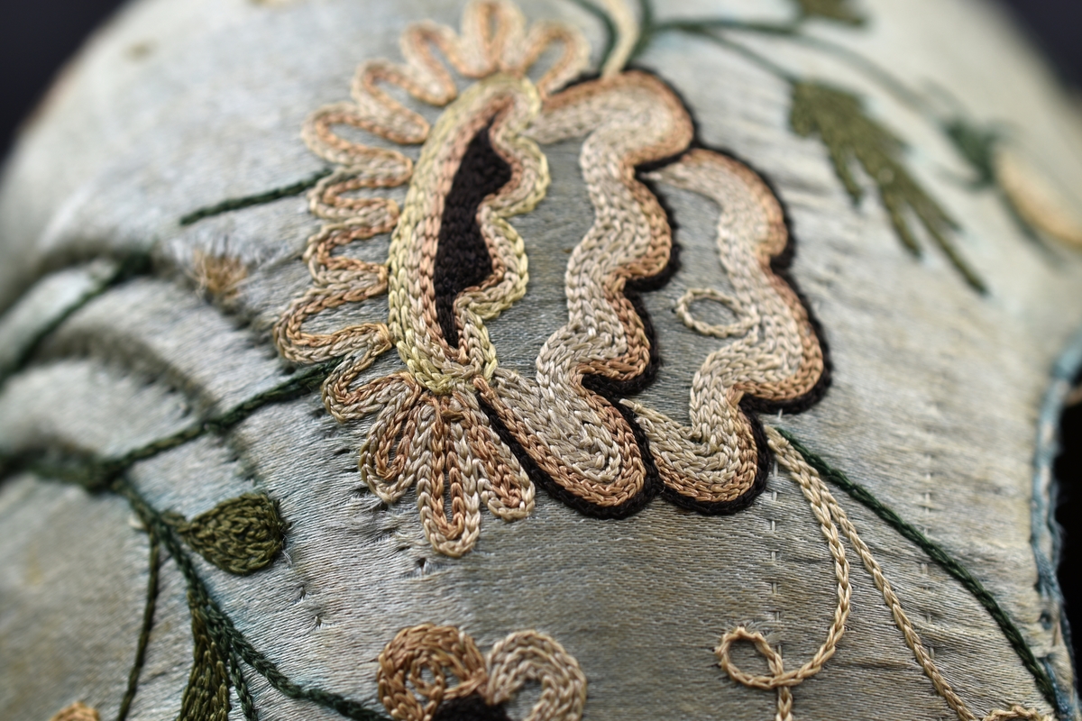 Bindmössa av ljusblått siden dekorerad med broderade blommor och blad. Foder i oblekt linnetyg.