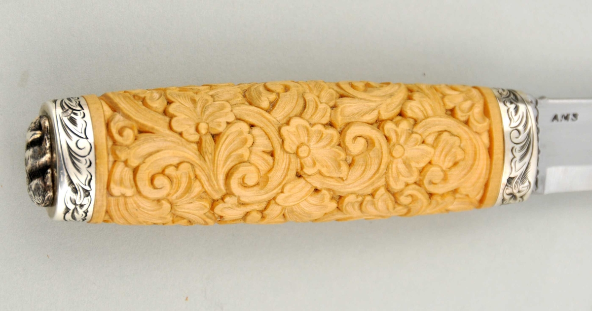 Kniv med utskore skaft og gravera holkar, på toppen ein støypt hund.
Slire av horn, utskoren på begge sider, holkar av sylv som er gravera.