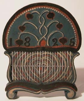 Bandvävsked av trä. Halvrunt, högt överstycke med ena sidan dekorerad med slingor samt ett årtal i relief och den andra sidan har en krona samt initaialer inom en ram och under glas. Vävskeden är målad i blått, rött, gult, vitt.