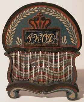 Bandvävsked av trä. Halvrunt, högt överstycke med ena sidan dekorerad med slingor samt ett årtal i relief och den andra sidan har en krona samt initaialer inom en ram och under glas. Vävskeden är målad i blått, rött, gult, vitt.