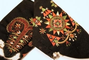 Svarta vinterkyrkvantar. Skinnfodrade med renskinn. Broderi utfört i stjälkstygn, plattsöm och flätsöm med silkegarn i grönt, brunt, vitt och gult. Inuti handen och på tummen öppningar kantade med mjukt vitt skinn. Öppningarna är till för kunna bläddra i psalmboken.