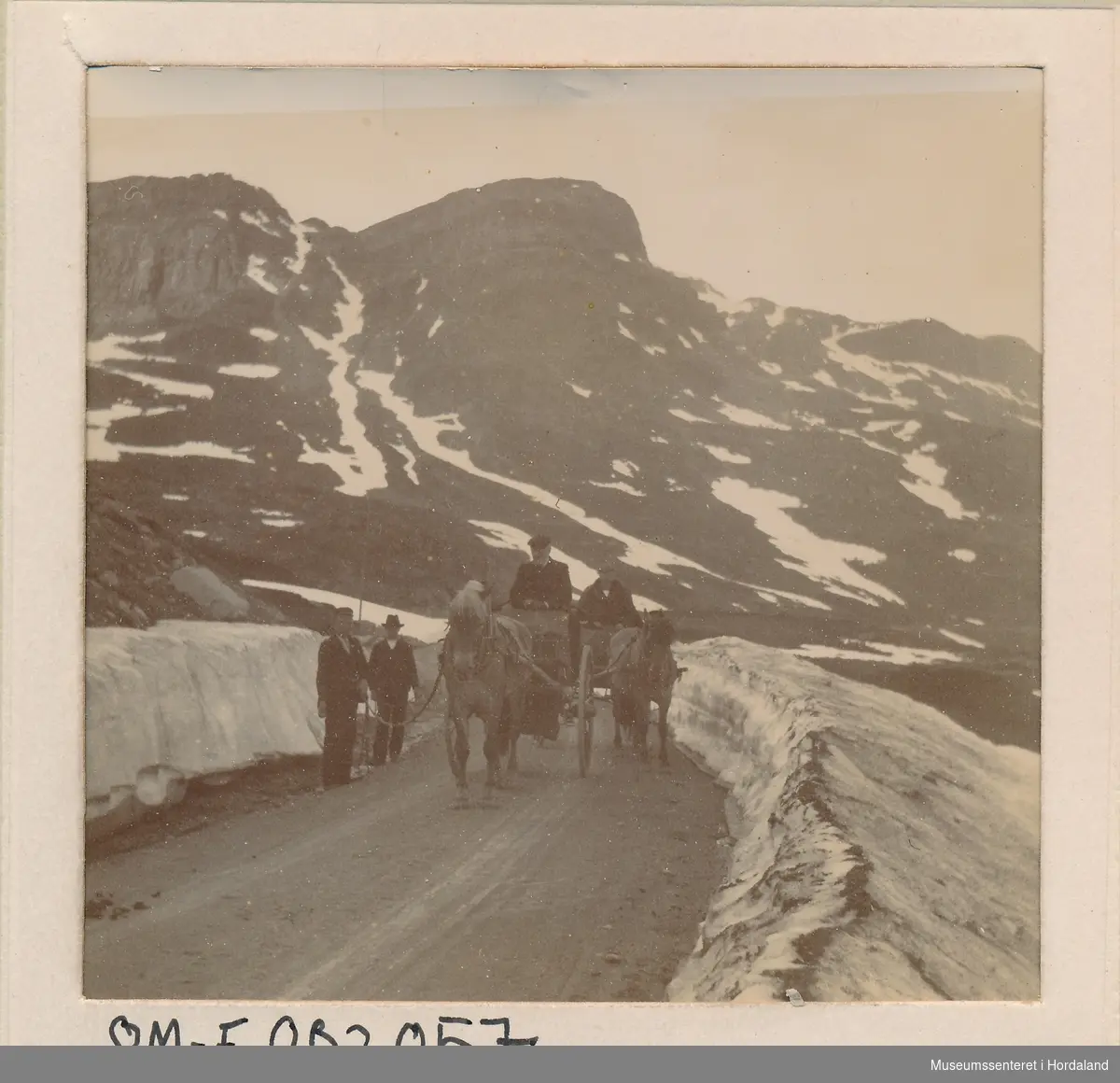 amatørfotografi av vegstrekning over fjell med brøytekantar, to hestar med vogn og kusk og to menn attmed