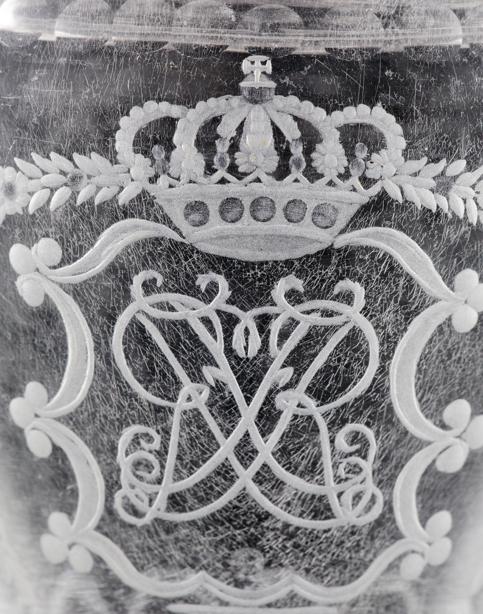 Pokal av glas, med lock. 
Slipat och graverat mönster med kungligt spegelmonogram: "FR"* i kartusch under krona. Invikt kant på foten.

*"FR" (Fredericus Rex= Fredrik I av Sverige, reg. 1720-1751).