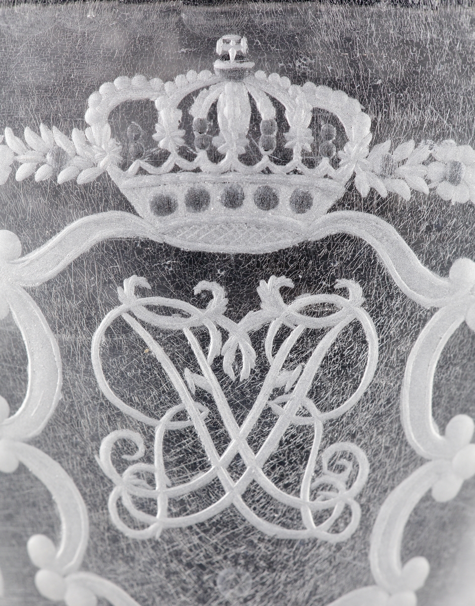 Pokal av glas, med lock. 
Slipat och graverat mönster med kungligt spegelmonogram: "FR"* i kartusch under krona.

*"FR" (Fredericus Rex= Fredrik I av Sverige, reg. 1720-1751).