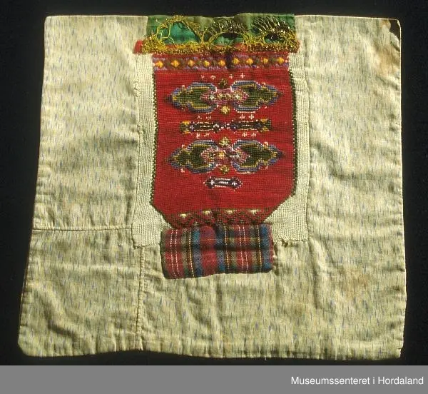 Bringeklut brodert i raudt med blå, grøn, gul, grøn, kvit, rosa og lilla. Gullfarga perler til dekor nokre stader, mange er knuste.  Grøn silkekvarde øvst med gullband i metall. Dette er sydd oppå eit raudt, smalt band. Rutete stoffbit sydd på under nederste broderte rad (Truleg på grunn av djup utringing på drakt). Montert på eit naturfarga, mønstra klede.