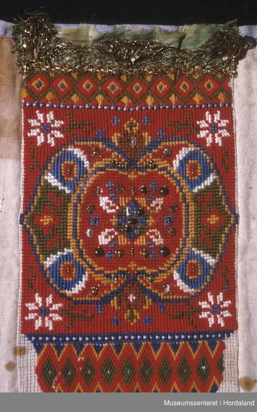 Bringeklut brodert på stramei med kross-sting og dragesaum. Raud med blått, gult, grønt og gull. Festa på eit kvitt stoff. Stoffet har ein stor flekk og nokre små. Oppe på baksida er montert på eit stripete stoff som går eit lite stykke ned på framsida. Grønt silkeband oppe med gullfarga metallkronekvarde sydd utanpå. Store og små perler sydd på broderimønsteret, nokre av desse er knuste.