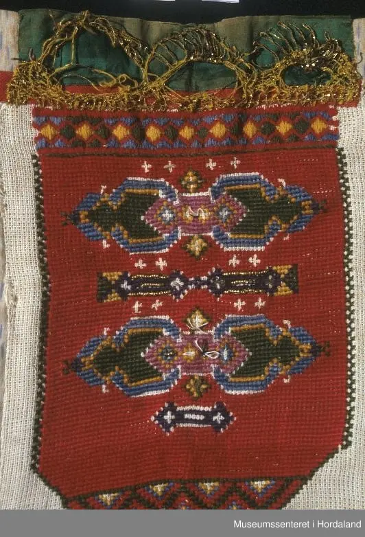 Bringeklut brodert i raudt med blå, grøn, gul, grøn, kvit, rosa og lilla. Gullfarga perler til dekor nokre stader, mange er knuste.  Grøn silkekvarde øvst med gullband i metall. Dette er sydd oppå eit raudt, smalt band. Rutete stoffbit sydd på under nederste broderte rad (Truleg på grunn av djup utringing på drakt). Montert på eit naturfarga, mønstra klede.