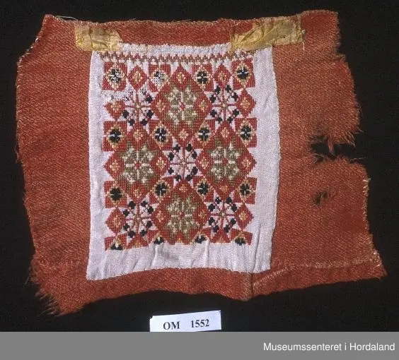 Bringeklut med kross-stingbroderi i raudt, grønt, gult, blått på kvit lin. Raudt er hovedfargen. 8-bladroser i mønsteret. Montert på eit raudt ullstoff. Restar etter silkekvarding oppe.