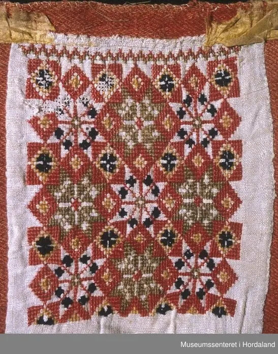 Bringeklut med kross-stingbroderi i raudt, grønt, gult, blått på kvit lin. Raudt er hovedfargen. 8-bladroser i mønsteret. Montert på eit raudt ullstoff. Restar etter silkekvarding oppe.