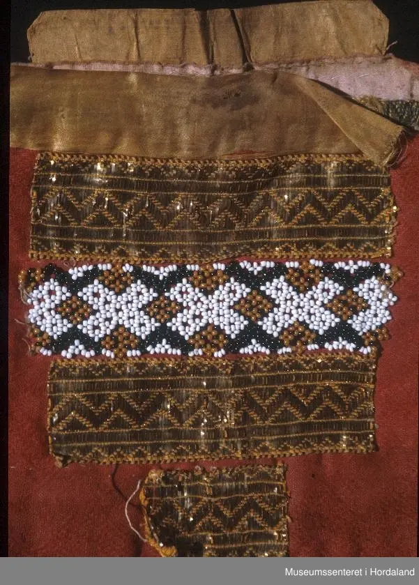 Bringeklut samansett av border på tvers, montert på raud ulldamask. Øvst ein gul silkekvarde montert utanpå ein krokakvarde. Bringekluten er forlenga i høgda frå baksida, og har eit silkeband i toppen som kjem over det andre. Bordene er to metallband med perlenettbord mellom. Nedst ein liten bit av same gullband. Mørkeblått klede til bakstoff.