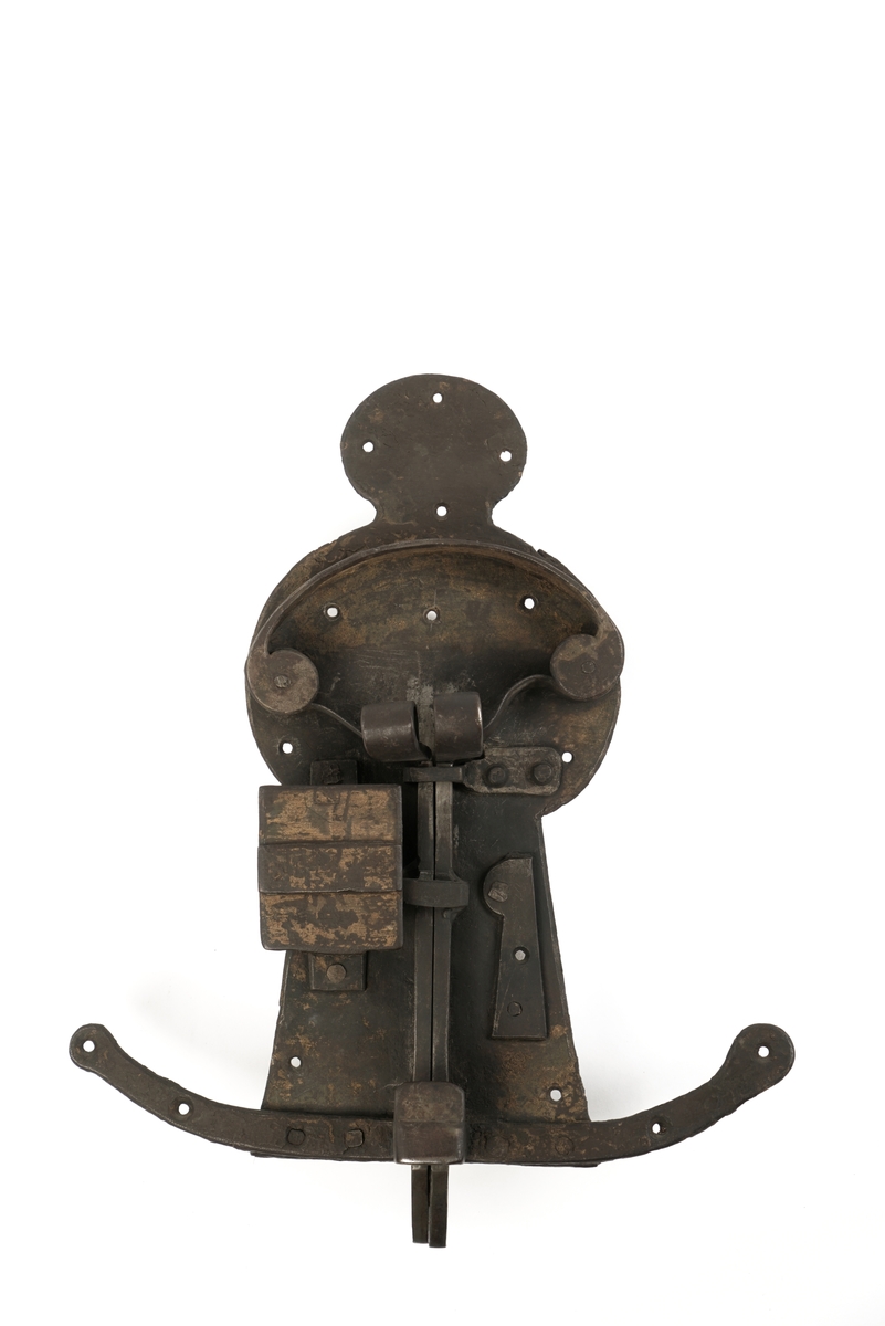 Dørlås av jern. Formet som en person, som står på en meie. Grov låsemekanisme med en hendel som gjør at låsebolten kan skyves til side, og dermed åpne døren.