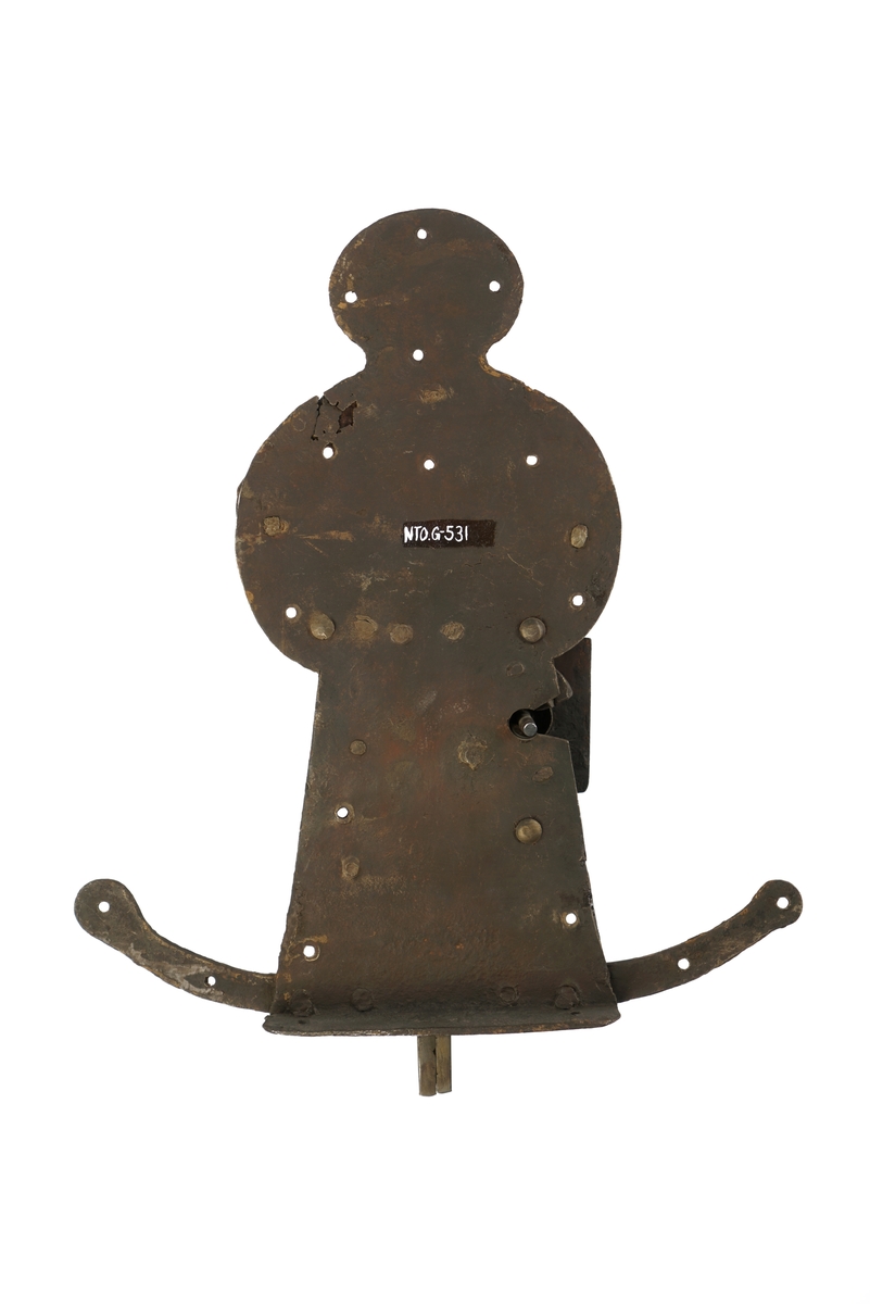 Dørlås av jern. Formet som en person, som står på en meie. Grov låsemekanisme med en hendel som gjør at låsebolten kan skyves til side, og dermed åpne døren.