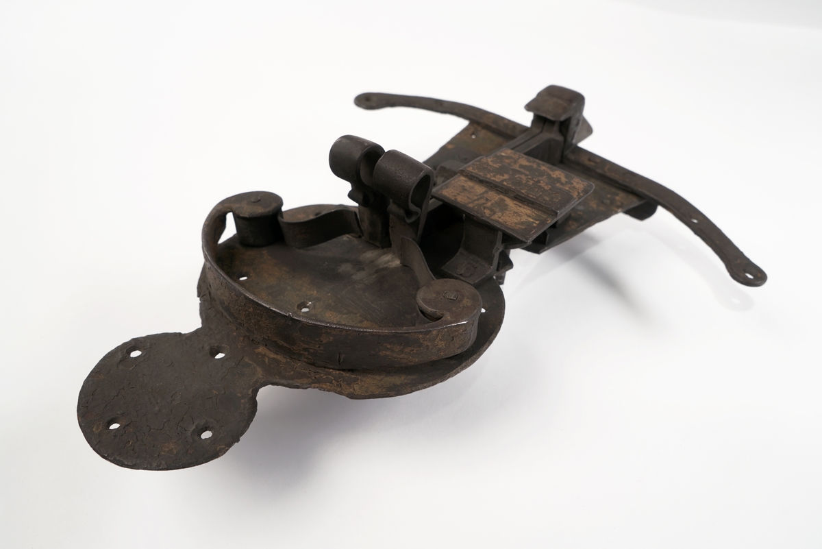 Dørlås av jern. Formet som en person, som står på en meie. Grov låsemekanisme med en hendel som gjør at låsebolten kan skyves til side, og dermed åpne døren.