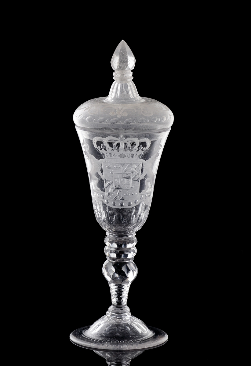 Pokal av glas, med lock.
Försedd med graverad dekor i form av Sveriges riksvapen* under kunglig krona, m.m.

*Såsom det var utformat under Fredrik I:s tid (reg. 1720-1751).
