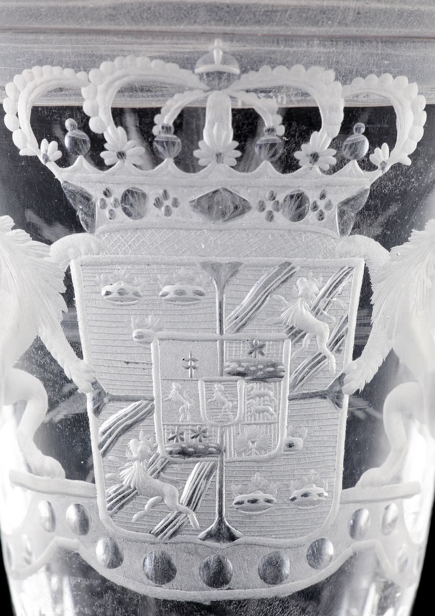 Pokal av glas, med lock.
Försedd med graverad dekor i form av Sveriges riksvapen* under kunglig krona, m.m.

*Såsom det var utformat under Fredrik I:s tid (reg. 1720-1751).