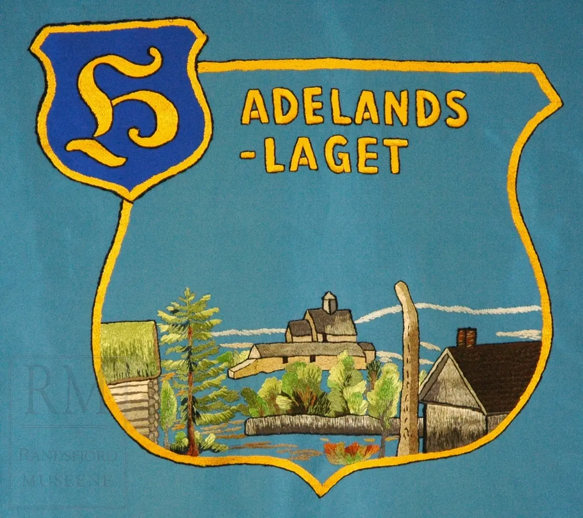 Fane; motiv og skrift på blå bunn. Skrift og possement i gull. Bæresele. Tre spyd. Brodert motiv på fremside: Utsikt fra Halvdanshaugen på Hadeland Folkemuseum, dynnasteinen i forgrunn, Tingalstad gamle kirke i bakgrunn. Bakside: Mariakirken og Nikolaikirken på Granavollen