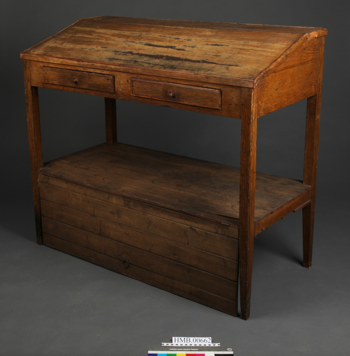 Rektangulært med skrånende skriveplate. To skuffer i front. Høye bein. Omtrent midt på beina er en hylleplate hvor det er hengslet en skapdør med en ring til håndtak. Skapdøren er hengslet i overkant og trekkes opp for å åpne.