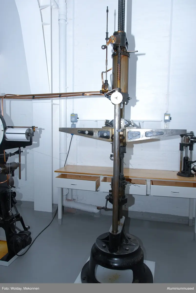 Strekkprøvemaskin for metaller.  Leverandør: Alfred J. Amsler & Co. Schweiz.  Datering: ca. 1925-1966. Teknikk: Hovedenhetene er støpt i stål og delvis maskinert.  Enkelte deler er fremstilt av valset stål og hydrauliske overføringer er kobber-rør.
Form: To stk. maskinenheter.  En betjeningsenhet og en strekkenhet som er forbundet med hydrauliske rør.
Tilstand: Maskinen er operativ, men har hydrauliske lekasjer.
Utfyllende opplysninger:
Betjeningsenheten består av en hydraulisk pumpe som via 2 stk. reguleringsventiler leverer olje til sylinderen for pendelmanometeret og til hovedsylinderen på toppen av strekkenheten.  Strekkprøvene plasseres i klemmebakkene på strekkenheten, blyanten settes på papirrullen og høyre kran på betjeningsenheten åpnes.  Etterhvert som trykket økes blir prøven strukket helt til brudd.  Under strekket slår pendelen ut tilsvarende trykket i hovedsylinderen og strekk-kraften kan til enhver tid avleses på skala-skiven.  Via et snorsystem fra overbjelken hvor øvre klemmbakke sitter, vil strekkforløpet bli avtegnet på en papir-rull på betjeningsenheten og strekkforløpet blir avtegnet slik at hele strekkforløpet og flytgrensen kan avleses.  Ved bruk avstenges hovedkranen umiddelbart og da vil den sorte pila umiddelbart gå tilbake til null-posisjon., men den røde pila blir stående igjen og viser bruddkraften.  Med den venstre kranen slippes hovedsylinderen ned til utgangsposisjon igjen.  Tykkelse og bredde på prøvestaven blir målt før prøvetaking og staven blir også risset med tynne paralelle strekker med 10 mm mellomrom.  Se risseapparat HAM.79.  Etter brudd blir prøvestaven satt sammen igjen og målt over en lengde på 5 riss med bruddet ca. midt i mellom. Den nye lengden i forhold til 50 mm viser forlengensen som blir utregnet i prosent.  Bruddfastheten er avlest bruddkraft delt på tverrsnitt.  Flytegrensen avleses av det tegende diagrammet på papir-rullen pluss 0,2%.  Maskinen kan maks. yte 10 tonn, men pga. unøyaktig avlesing ved tynne prøver blir pendelmanometeret innstilt ettter forventet bruddstyrke.  Maskinen har 4 innstillnger: Maks.: 1000 kg: Nest nederste hull. Uten lodd. Maks.: 2000 kg: Nest øverste hull. Uten lodd. Maks.: 5000 kg: Nederste hull.  Med Lodd. Maks.: 10000 kg.: Nest øverste hull. Med lodd.
Maskinen står i dag 08.01.2007 i psosisjon, 5000 kg.  Maskinen kan også brukes til trykkprøving, f.eks. sveiseprøver, ved å plassere prøven på rullebakkene oppe på strekk-bommen.  22.01.2008 Kjell Grette.
