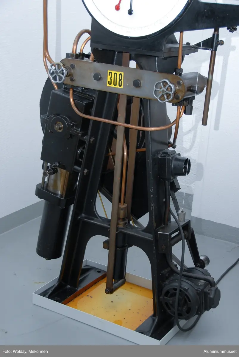 Strekkprøvemaskin for metaller.  Leverandør: Alfred J. Amsler & Co. Schweiz.  Datering: ca. 1925-1966. Teknikk: Hovedenhetene er støpt i stål og delvis maskinert.  Enkelte deler er fremstilt av valset stål og hydrauliske overføringer er kobber-rør.
Form: To stk. maskinenheter.  En betjeningsenhet og en strekkenhet som er forbundet med hydrauliske rør.
Tilstand: Maskinen er operativ, men har hydrauliske lekasjer.
Utfyllende opplysninger:
Betjeningsenheten består av en hydraulisk pumpe som via 2 stk. reguleringsventiler leverer olje til sylinderen for pendelmanometeret og til hovedsylinderen på toppen av strekkenheten.  Strekkprøvene plasseres i klemmebakkene på strekkenheten, blyanten settes på papirrullen og høyre kran på betjeningsenheten åpnes.  Etterhvert som trykket økes blir prøven strukket helt til brudd.  Under strekket slår pendelen ut tilsvarende trykket i hovedsylinderen og strekk-kraften kan til enhver tid avleses på skala-skiven.  Via et snorsystem fra overbjelken hvor øvre klemmbakke sitter, vil strekkforløpet bli avtegnet på en papir-rull på betjeningsenheten og strekkforløpet blir avtegnet slik at hele strekkforløpet og flytgrensen kan avleses.  Ved bruk avstenges hovedkranen umiddelbart og da vil den sorte pila umiddelbart gå tilbake til null-posisjon., men den røde pila blir stående igjen og viser bruddkraften.  Med den venstre kranen slippes hovedsylinderen ned til utgangsposisjon igjen.  Tykkelse og bredde på prøvestaven blir målt før prøvetaking og staven blir også risset med tynne paralelle strekker med 10 mm mellomrom.  Se risseapparat HAM.79.  Etter brudd blir prøvestaven satt sammen igjen og målt over en lengde på 5 riss med bruddet ca. midt i mellom. Den nye lengden i forhold til 50 mm viser forlengensen som blir utregnet i prosent.  Bruddfastheten er avlest bruddkraft delt på tverrsnitt.  Flytegrensen avleses av det tegende diagrammet på papir-rullen pluss 0,2%.  Maskinen kan maks. yte 10 tonn, men pga. unøyaktig avlesing ved tynne prøver blir pendelmanometeret innstilt ettter forventet bruddstyrke.  Maskinen har 4 innstillnger: Maks.: 1000 kg: Nest nederste hull. Uten lodd. Maks.: 2000 kg: Nest øverste hull. Uten lodd. Maks.: 5000 kg: Nederste hull.  Med Lodd. Maks.: 10000 kg.: Nest øverste hull. Med lodd.
Maskinen står i dag 08.01.2007 i psosisjon, 5000 kg.  Maskinen kan også brukes til trykkprøving, f.eks. sveiseprøver, ved å plassere prøven på rullebakkene oppe på strekk-bommen.  22.01.2008 Kjell Grette.