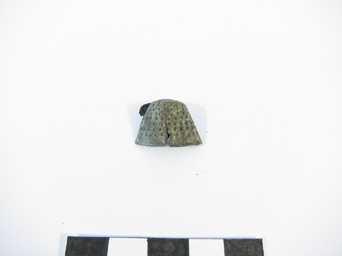 Svagt konisk fingerborg av brons med relativt glesa gropar. Nedtill är en slät rand med fabriksstämpel: H intryckt (se bild).