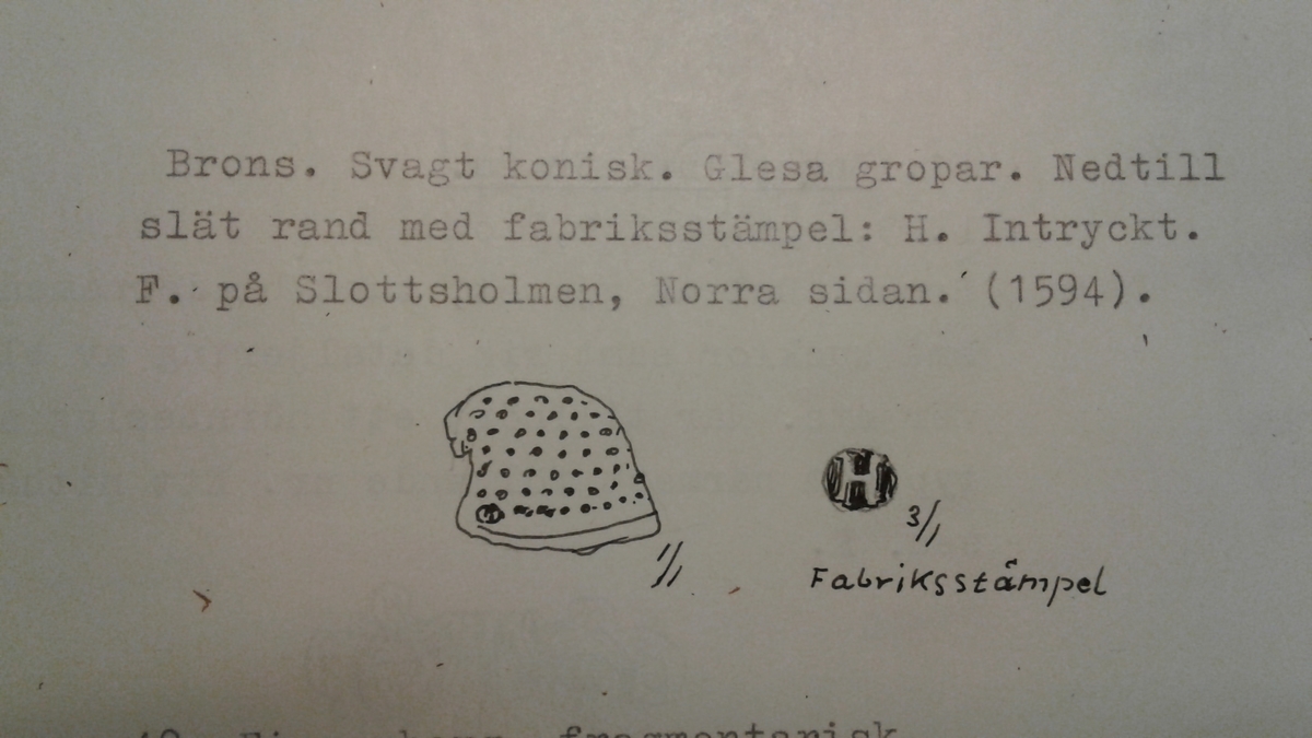 Svagt konisk fingerborg av brons med relativt glesa gropar. Nedtill är en slät rand med fabriksstämpel: H intryckt (se bild).