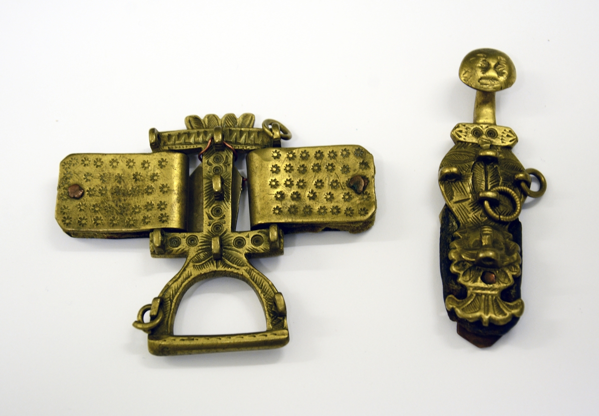 Slirehempe til belte og beltelås i messing. Fra protokollen: Bæltebeslag for tollekniver, av messing. Selve beslaget bestaar av a) 1 sprote (stolpelås eller beltelås) med paasatte 4-5 ringer og ornamenter i form av to masker.
b) En stolpe (slirehempe) med løkke paa. Fra stolpen gaar 2 par tvertagger, som gir plads til rembeslagene. Disse, der bestaar av 2 dobbeltbiøede plater ornerte med stjernepunkter, gaar rundt mellem tvertaggerne, og omslutter skindremmen som bokpermer. Beslaget har ringe og ornamenter.