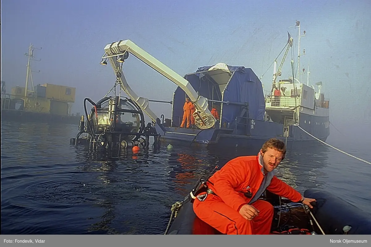 En mann kjører bort fra dykkerskipet "Strilborg" i en liten gummibåt.  Strilborg ble senere omdøpt til "Deepsea Inspector", og er moderskipet til miniubåten "Check Mate" som henger i kranbommen i akterenden av skipet.