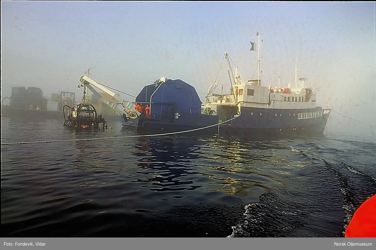 En mann kjører bort fra dykkerskipet "Strilborg" i en liten gummibåt.  Strilborg ble senere omdøpt til "Deepsea Inspector", og er moderskipet til miniubåten "Check Mate" som henger i kranbommen i akterenden av skipet.