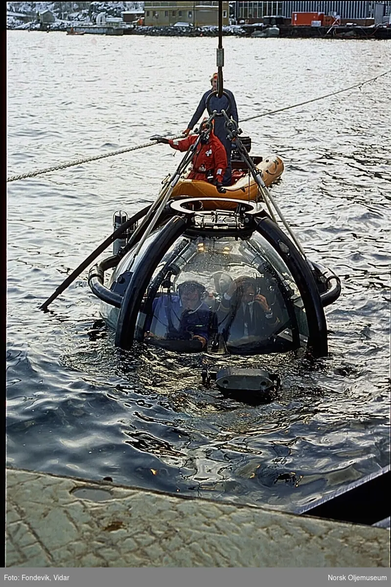 Miniubåten "Check Mate" heises opp eller er på vei ned i sjøen i  Gravdalsbukta. To personer bemanner ubåten, mens to andre følger med i en liten gummibåt like ved.  Den ene mannen i gummibåten er kledd i overlevelsesdrakt.  NUI ligger i bakgrunnen.