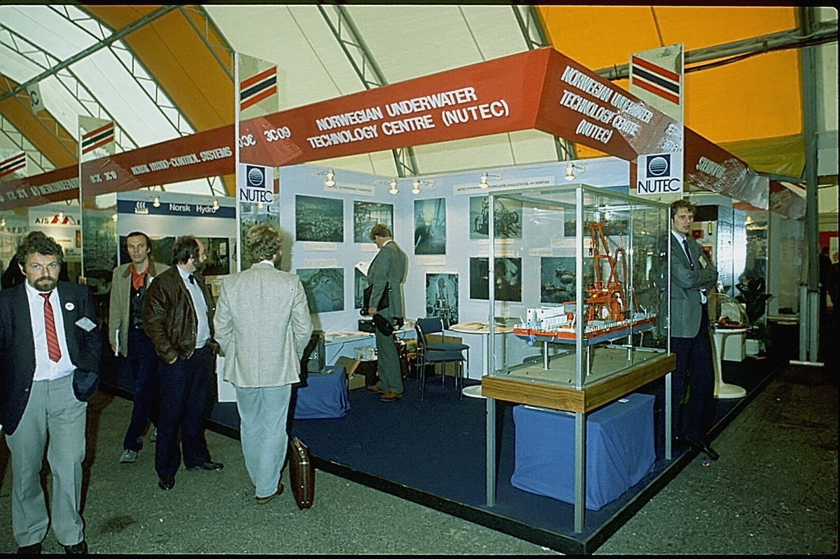 NUTEC sin stand på en messe. En person betjener standen.  En modell av "NUTEC Fjordbase" er plassert i en monter.