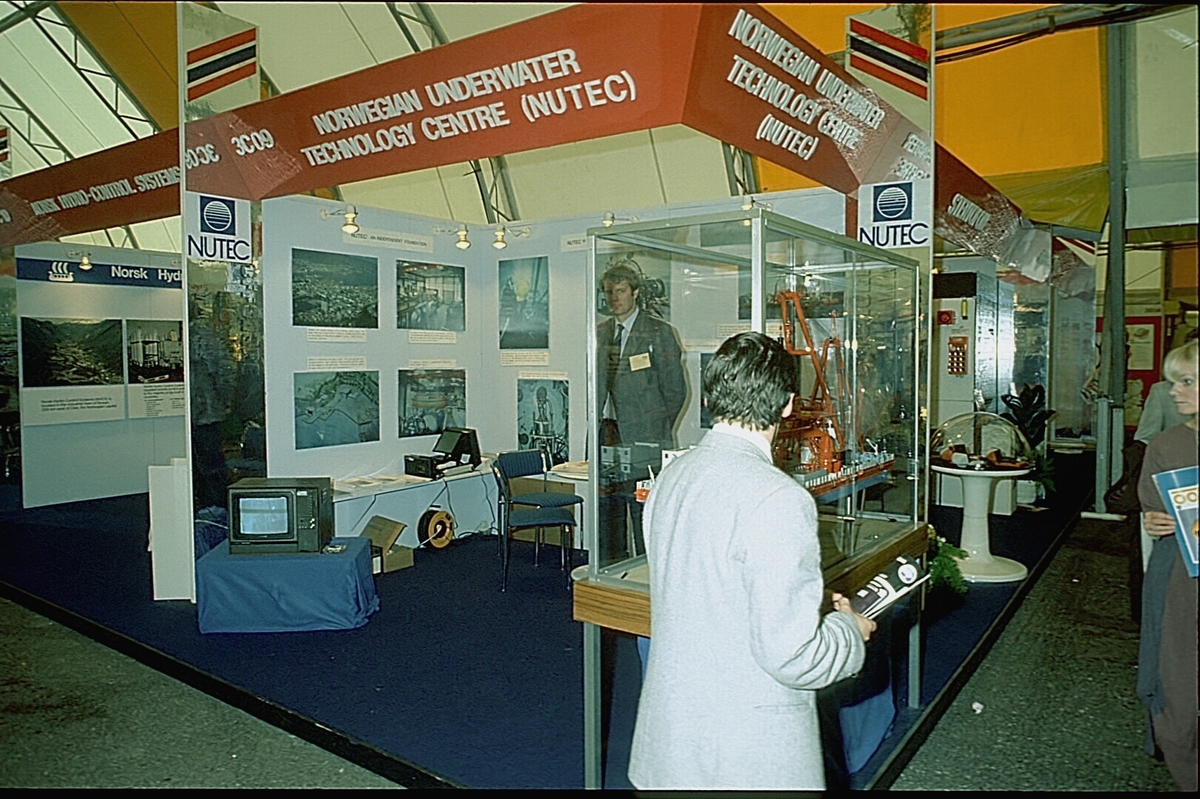 NUTEC sin stand på en messe. En person betjener standen.  En modell av "NUTEC Fjordbase" er plassert i en monter.