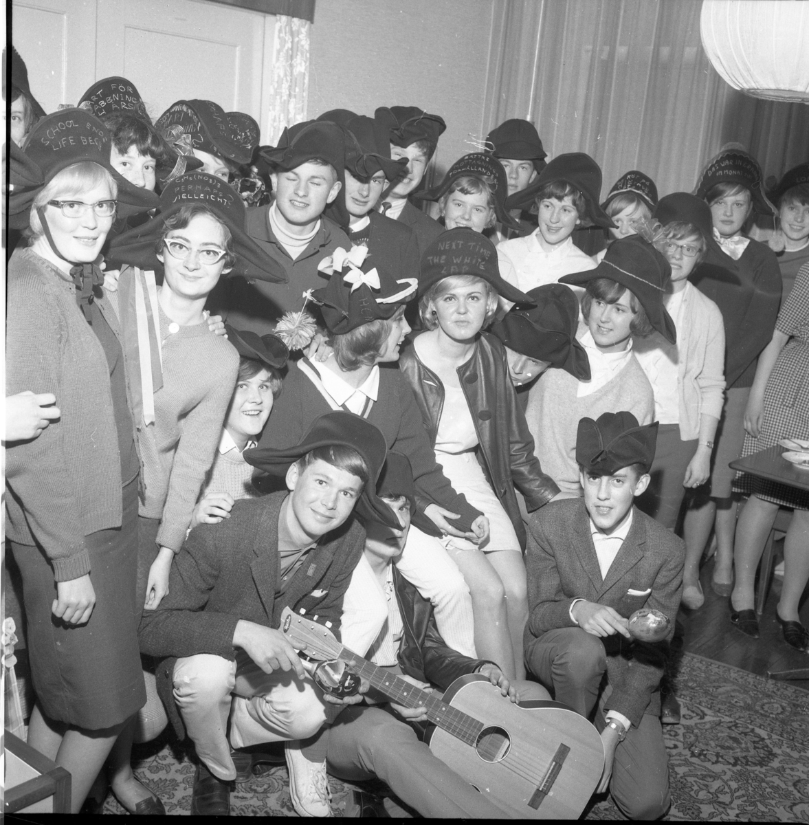 En grupp abiturienter med hattar, märkta på olika sätt, 1964. Längst fram sitter Bo Palm, okänd samt Thomas Sonesson. Andra ledet: Berit Larsson, Zolie Axelsson, Ewa Heldebrandt, Susanne Arding, Ingrid Lööf, okänd kille samt Lena Simonsson. Nästa leda skymtar till vänster Berit Johansson och Iris Gustafsson, därnäst Morgan Karlsson, Nils-Erik Olofsson, okänd, Ann-Christina Haglund, Thorsten Rydberg, Gullan Hagström, Britt-Marie Karlsson.