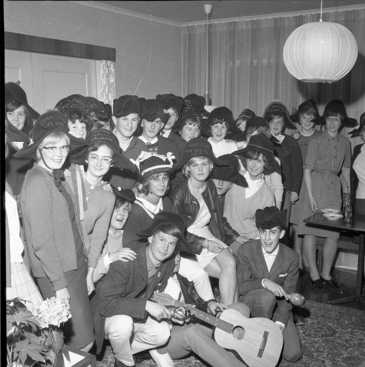 En grupp abiturienter med hattar, märkta på olika sätt, 1964. Längst fram sitter Bo Palm, okänd samt Thomas Sonesson. Andra ledet: Berit Larsson, Zolie Axelsson, Ewa Heldebrandt, Susanne Arding, Ingrid Lööf, okänd kille samt Lena Simonsson. Nästa led skymtar okänd, Berit Johansson och Iris Gustafsson, därnäst Morgan Karlsson, Nils-Erik Olofsson, okänd, Ann-Christina Haglund, Gullan Hagström, Britt-Marie Karlsson. I gruppen finns också Gunborg Svensson från Bunn.