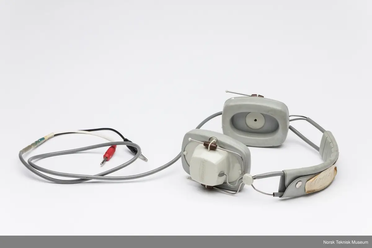 Hodetelefon med ledning og to plugger. Papirlapp festet til ene klokken med en tråd og tapebit på bøyle.