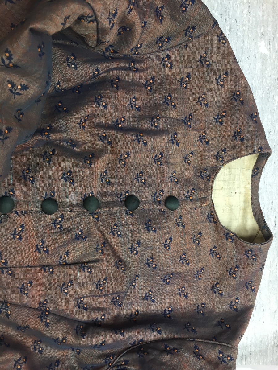 Kjole sammensydd av liv og skjørt med belte i livet med sløyfe i samme stoff. Halvull verkenstoff med trykte blomster. Sydd i Sørum i Akershus ca 1860 og senere brukt på Hamar. Skjørtet har mye håndsøm men vi ser også maskinsøm. Skjørtet er fóret med en skoning av lin i nedre kant. Det er også festet et grønt smalt bånd som slitekant. Overdelen er fóret med ufarget linstoff. Svart fløyelsbånd er festet 1,5 cm fra ermekanten.